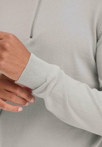 Lys grå glidelås hettegenser med ribbestrikkede mansjetter, myk tekstur og tettsittende design synlig på en persons arm og hånd.