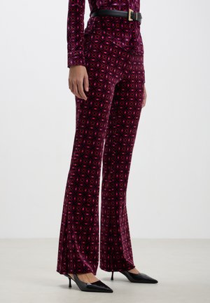 Pantaloni a palazzo in velluto di un profondo viola con un motivo geometrico a diamante in rosa e blu. Presentano una vita aderente e una cintura nera.