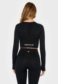 Černý sportovní crop top s dlouhými rukávy, texturovaná tkanina a otevřený design na zádech. Ladící legíny s vysokým pasem, také černé, s detailním logem.