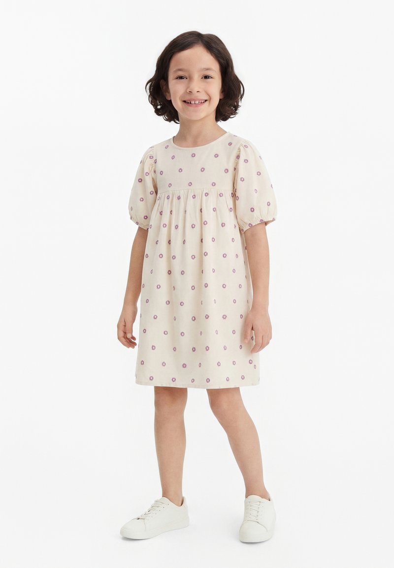 Fată tânără cu păr scurt și creț zâmbitoare, purtând o rochie crem cu mici modele cu flori roz și adidași albi, stând pe un fundal alb.