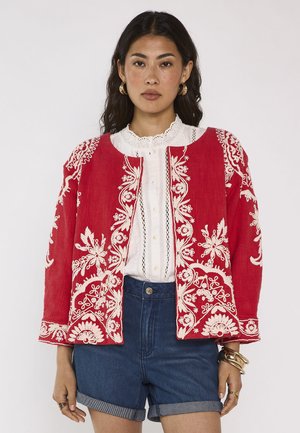 Donna con lunghi capelli scuri indossa una giacca rossa con ricamo floreale bianco, una camicetta di pizzo bianca, pantaloncini di jeans blu, orecchini a cerchio dorati e braccialetti.