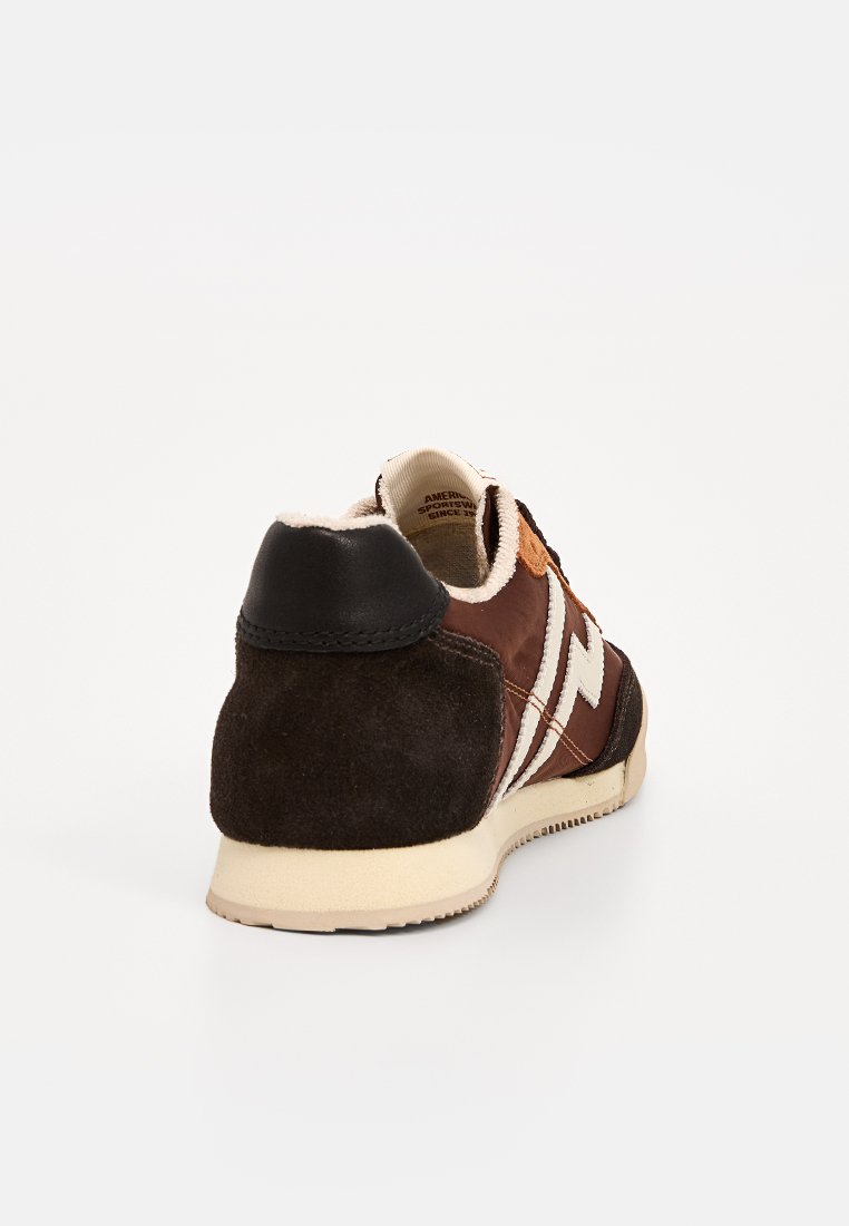 Braune Wildleder- und Leder-Sneaker mit einem schwarzen Fersenbereich, beiger Sohle und weißen Zickzackakzenten. Die Rückansicht zeigt die Nähdetails.