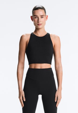 Žena s vlasy staženými dozadu, oblečená v černém crop topu bez rukávů a černých legínách s vysokým pasem, stojící před jednobarevným světlým pozadím.