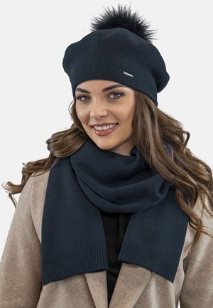 Beret negru tricotat cu un pom-pom din blană sintetică în vârf, completat cu o eșarfă neagră asortată, evidențiind o textură moale și un design elegant.