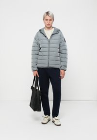 Grå pufferjakke med horisontale syninger, lynlåslukning og hætte; marineblå bukser; holder en sort tote bag; hvide sneakers med mørke detaljer.