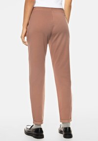 Pantalons stretch marron clair avec une coupe ajustée, ourlets à revers et taille élastique. Portés avec des chaussures noires et des chaussettes grises.