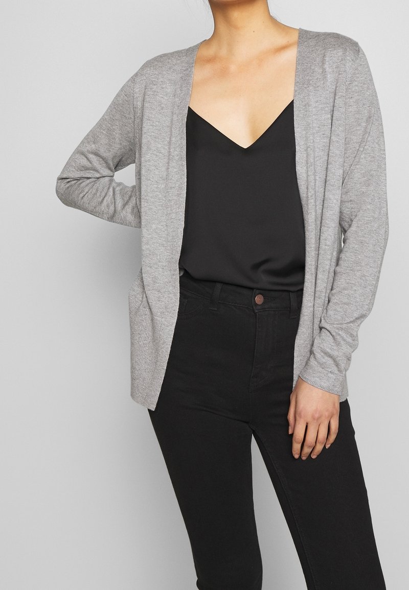 Cardigan en maille gris clair avec ouverture sur le devant, manches longues et deux poches avant, porté sur un haut noir à col en V et un pantalon noir taille haute.