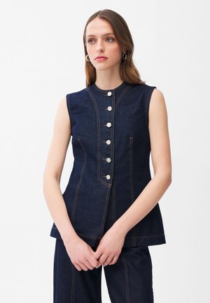 Giovane donna che indossa un top senza maniche in denim scuro con bottoni e pantaloni abbinati, in piedi davanti a uno sfondo bianco uniforme.