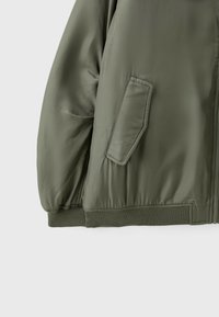 Gros plan d'une manche de veste vert olive et du bas devant avec une poche à rabat inclinée, un poignet et un ourlet élastiques.