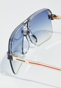 Ochelari de soare cu lentile mari de gradient albastru, margini gri translucide, accente metalice aurii și o dungă roșie pe brațul purtând inscripția "CARRERA."