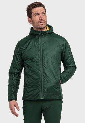 Mann trägt eine dunkelgrüne gesteppte Kapuzenjacke mit passenden Hosen, schaut nach oben und links mit entspannten Händen in den Taschen.
