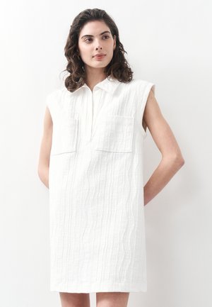 Kiharaliste juustega naine kannab kaelusega ja kahe rinnataskuga varrukateta valget tekstuurset kleiti ning seisab ühevärvilise valge tausta ees.