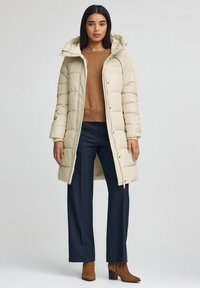 Beżowa pikowana kurtka z kapturem, z przodu zapinana na zamek błyskawiczny i zatrzaski, zestawiona z brązowym swetrem i granatowymi spodniami.