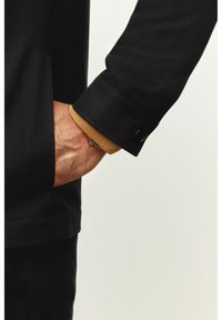 Sort jakke med tekstureret stof, med en brun ribbet manchet. Hånd placeret i en sidelomme, fremhævet af en knapdetalje.