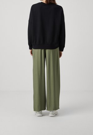 Osoba ubrana w czarną bluzkę z długim rękawem, luźne oliwkowozielone spodnie i białe trampki, stojąca tyłem na jednolitym tle.