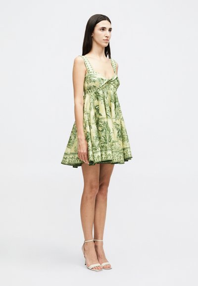 Γυναίκα με μακριά σκοτεινά μαλλιά που φοράει αμάνικο μίνι φόρεμα με πράσινα και μπεζ λουλούδια και μπεζ ψηλοτάκουνα σανδάλια, στέκεται μπροστά σε λευκό φόντο.