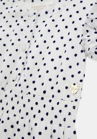 Weisses Kleid mit marineblauen Punkten, kurzen Rüschensäumen, gerafftem Bund und einem herzförmigen Metallschild mit "Guess"-Logo.