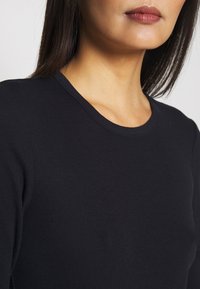 Musta ribbineulottu pitkähihainen paita pyöreällä pääntiellä. Kangas on sileää, siinä on hienovarainen tekstuuri ja vartalonmyötäinen istuvuus.