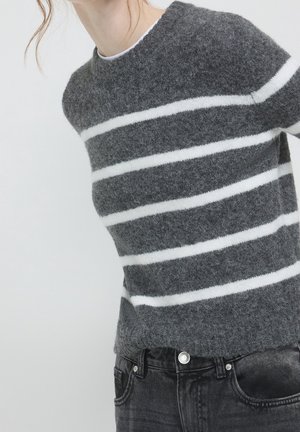 Personne portant un pull gris duveteux avec trois bandes horizontales blanches et un jean noir, sur un fond clair uni.