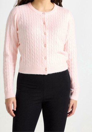 Kvinde iført en lyserød kabelstrikket cardigan med knapper, kombineret med sort, højtaljede bukser, stående foran en ensfarvet baggrund.