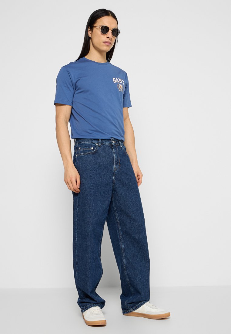 Blaues Baumwoll-T-Shirt mit Logo, kombiniert mit lässigen, dunkelblauen Jeans. Weiße Sneaker mit beige-farbigen Sohlen. Schwarze Sonnenbrille vervollständigt das Outfit.