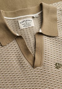 Polo en maille à motifs beige et blanc avec col et encolure en V, portant une étiquette Superdry Co et un logo brodé sur la poitrine.