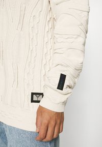 Sweter w kolorze kremowym, dzianinowy, z teksturowanymi wzorami w warkocze, ściągaczami przy mankietach oraz z naszywkami markowymi na rękawie i boku.