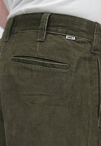 Olivnozeleni kordurojski hlače s detajli na zadnjem žepu, z majhno "OBEY" nalepko našito na pasu. Teksturirano, navpično rebrasto blago.