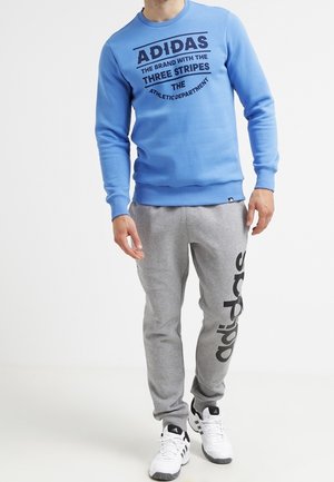 Mann trägt einen blauen Adidas-Sweatshirt mit schwarzem Schriftzug und graue Jogginghose mit großem schwarzem Adidas-Logo am linken Bein, kombiniert mit weißen Sneakers.