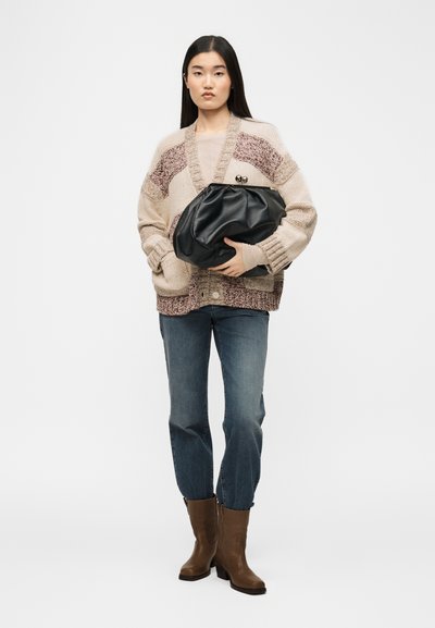 Γυναίκα που φοράει μπέζ και ροζ πλεκτή ζακέτα, μπλε τζιν, καφέ μπότες, κρατώντας μεγάλη μαύρη δερμάτινη τσάντα φάκελο, στέκεται σε λευκό φόντο.