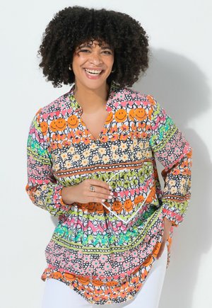 Kvinde med krøllet hår smiler, iført en farverig mønstret bluse med smilende ansigter og blomsterdesigns, holder klare briller.