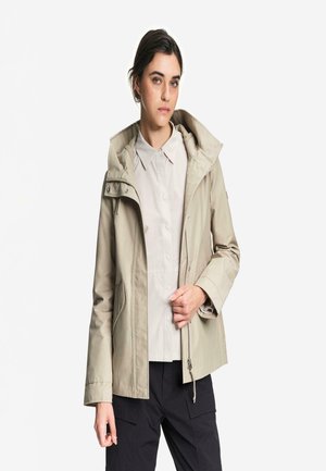 Mujer joven que lleva una chaqueta beige con capucha sobre una camisa abotonada y pantalones negros, de pie frente a un fondo blanco liso.