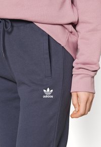 Granatowe spodnie dresowe z elastycznym paskiem, bocznymi kieszeniami i białym logo Adidas. Na górze znajduje się różowa bluza z ribbowanymi mankietami.