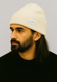Cream-colored ribbed beanie with a folded brim and a small blue brand label. Worn by a person with dark hair and a beard.