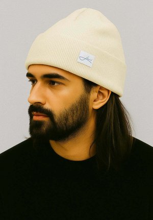 Cream-colored ribbed beanie with a folded brim and a small blue brand label. Worn by a person with dark hair and a beard.