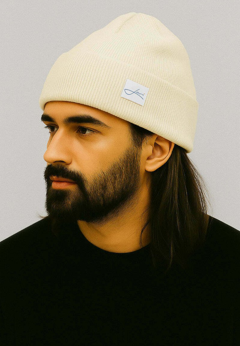 Cream-colored ribbed beanie with a folded brim and a small blue brand label. Worn by a person with dark hair and a beard.