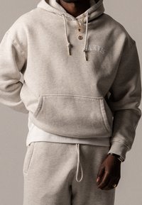 Mężczyzna w jasnobeżowej bluzie z kapturem i pasujących dresach, noszący złote łańcuchy, pierścionek i czarny zegarek na neutralnym tle.