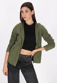 Oliwkowa bluza z kapturem na zamek, z długimi rękawami, wykonana z miękkiego polaru, w zestawie z dopasowaną czarną bluzką i czarnymi jeansami z wysokim stanem oraz paskiem.