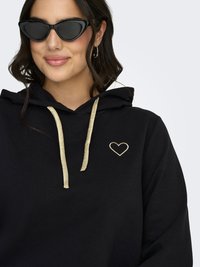 Nainen, joka käyttää mustia kissansilmä-aurinkolaseja ja mustaa hupullista paitaa, jossa on kultaiset nyörit ja pieni kullalla kirjailtu sydän rinnassa.