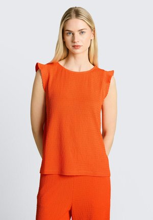 Femme blonde portant un haut sans manches texturé orange vif et un pantalon assorti, debout devant un fond gris clair.