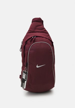 Vinrød Nike skuldertaske med frontlynlås lomme, hvid swoosh-logo og sort Nike mærkat hængende fra sidelåsen.