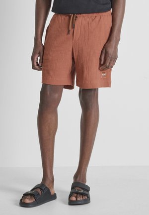 Man draagt roestkleurige, getextureerde korte broek met trekkoord en zwarte slides tegen een effen achtergrond.