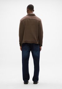 Giacca a collo marrone con una texture liscia, abbinata a jeans di denim scuri con due tasche posteriori e dettagli di cucitura chiara.