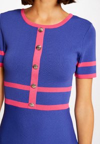 Abito lavorato a maglia di un blu reale con accenti a righe rosa e maniche corte. Presenta un collo tondo e sei bottoni decorativi argentati lungo la parte anteriore.