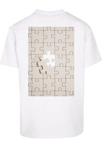 Hvid t-shirt med en grafik af et beige puslespil med én manglende brik. Designet er enkelt med klare konturer og en glat overflade.