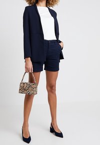 Laivaston sininen bleiseri valkoisen topin päällä, yhdistetty laivaston sinisiin shortseihin. Asusteena ruskean ja vaaleanruskean käärmekuvioinen käsilaukku ja laivaston siniset korkokengät.