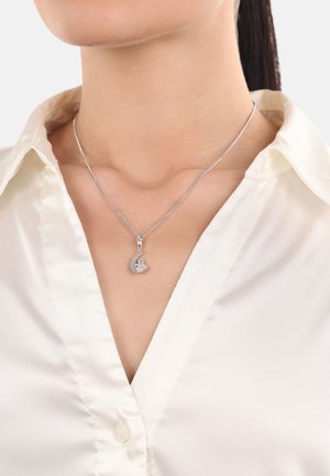 Silberne Halskette mit einem eleganten Anhänger, der ein kreisförmiges Design und ein kleines Dekorelement aufweist, getragen über einem weißen Satinhemd.