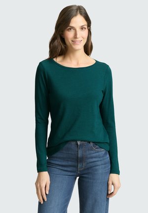 Femme aux longs cheveux bruns portant un haut à manches longues vert foncé et un jean bleu, souriante, debout devant un fond clair uni.