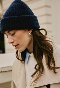 Granatowy, dziany beanie, beżowy płaszcz z czarnymi guzikami, jasnoniebieska koszula z kołnierzykiem, srebrne kolczyki w kształcie kół. Włosy delikatnie opadają na ramię.
