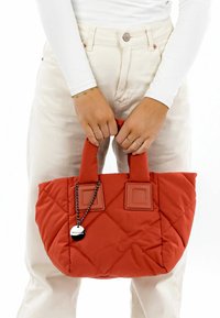 Quilted red handbag with two short handles, featuring square accents and a chain with a circular tag. The bag has a soft texture.
